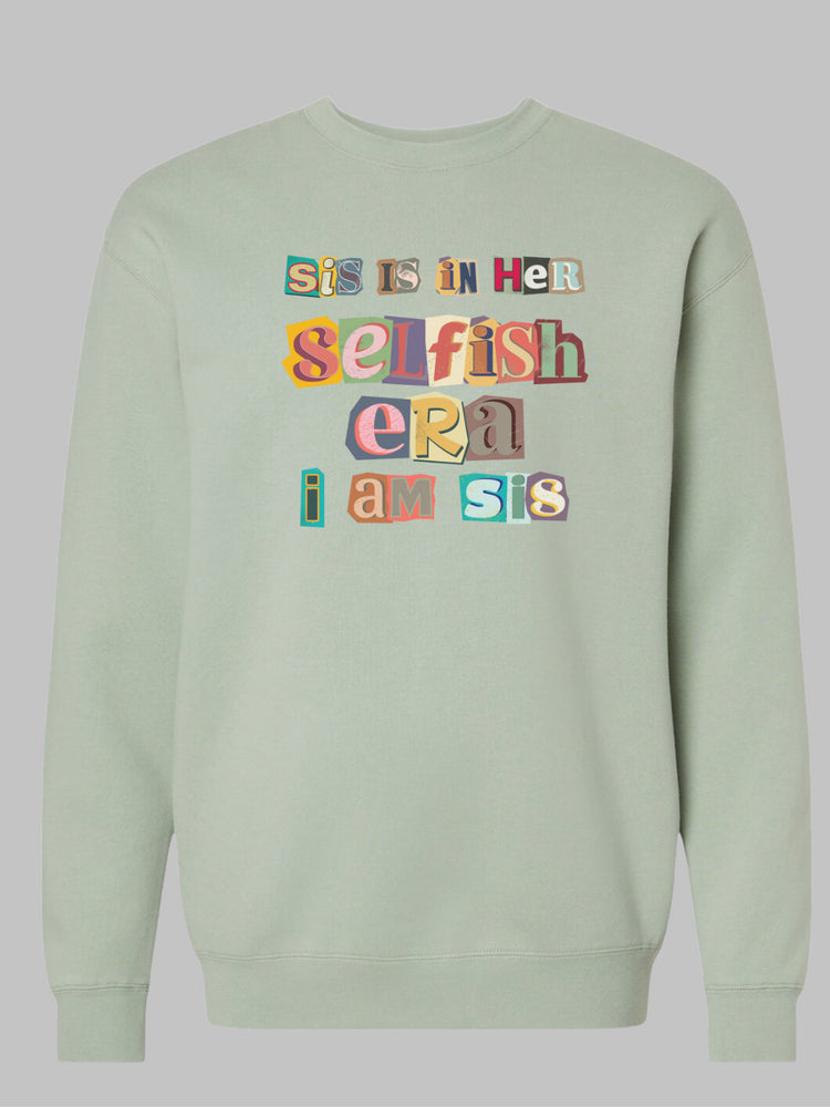 Image of a sage green -colored crewneck sweatshirt featuring the phrase 'Sis in her Selfish era. I am Sis' in a unique, multicolored ripped paper font. The text, appearing as if it's made from torn pieces of paper in various colors, creates a dynamic and artistic effect against the neutral tan background. This design conveys a message of self-empowerment and individuality, with the playful and creative font adding a touch of whimsy and boldness to the statement. Self love sweatshirt. self care sweatshirt