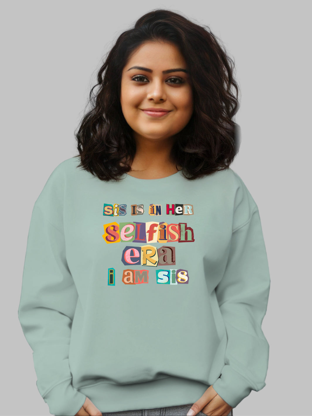 Image of a sage green crewneck sweatshirt featuring the phrase 'Sis in her Selfish era. I am Sis' in a unique, multicolored ripped paper font. The text, appearing as if it's made from torn pieces of paper in various colors, creates a dynamic and artistic effect against the neutral tan background. This design conveys a message of self-empowerment and individuality, with the playful and creative font adding a touch of whimsy and boldness to the statement. self love sweatshirt, self care crew neck sweatshirt