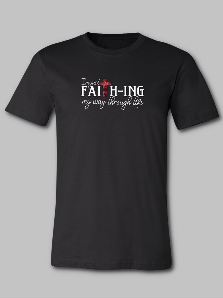 Image of a black unisex faith based crewneck t-shirt featuring the phrase 'Faith-ing It' in bold white font. Just above the text, there's a striking red rhinestone cross that adds a touch of sparkle and symbolism to the design. The contrast of the white text and red rhinestone against the black fabric creates a visually impactful statement of faith and perseverance.  Christian t-shirt