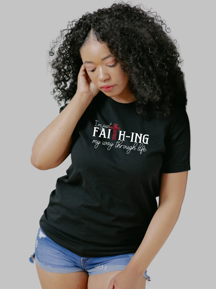 Image of a black unisex faith based crewneck t-shirt featuring the phrase 'Faith-ing It' in bold white font. Just above the text, there's a striking red rhinestone cross that adds a touch of sparkle and symbolism to the design. The contrast of the white text and red rhinestone against the black fabric creates a visually impactful statement of faith and perseverance.  Christian t-shirt